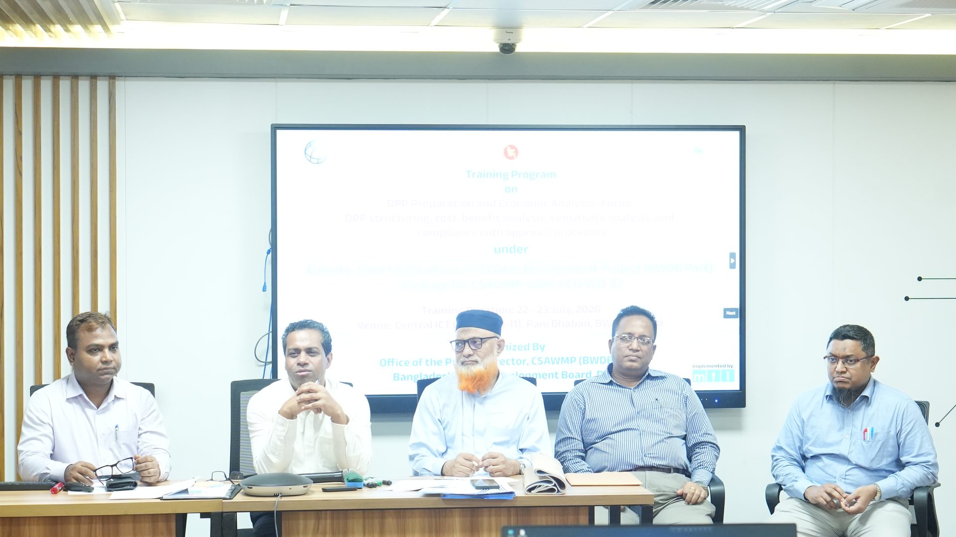 The Project Director addressing the DPP Preparation and Economic Analysis training from the head table, with the programme banner displayed on screen