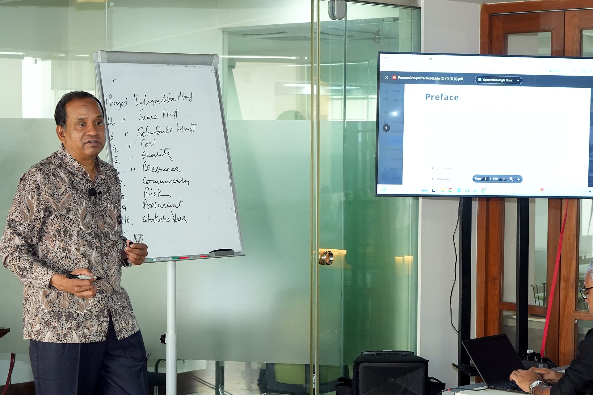 Trainer beside a flipchart listing the ten knowledge areas, with the PMI process groups guide on screen